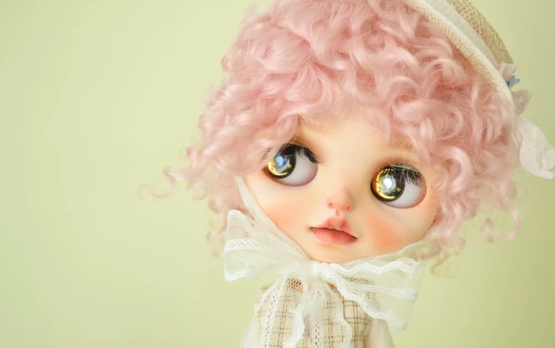 K&ouml;nnte beinhalten: Eine Puppe mit pinkfarbenen, lockigen Haaren, die einen wei&szlig;en Hut und eine wei&szlig;e Schleife aus Spitze tr&auml;gt. Die Puppe hat gro&szlig;e, blaue Augen mit goldenen Flecken.