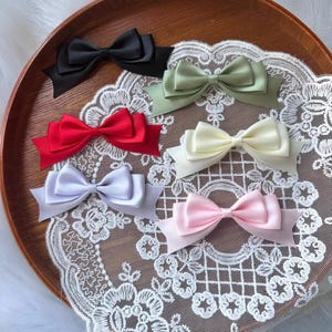 Può includere: Una collezione di sei fiocchi per capelli in raso nei colori nero, verde salvia, rosso, crema, grigio chiaro e rosa pallido. Ogni fiocco presenta un design a doppio strato con tessuto liscio e lucido e punte, disposto su un centrino di pizzo.
