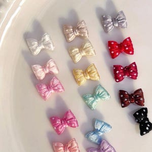 May include: A selection of small fabric bows in a range of colours, such as white, pink, red, yellow, and blue. Some bows have a polka dot design. These decorative bows are arranged on a white surface.