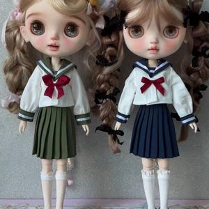 May include: Two dolls with large eyes and long hair styled in braids. Both wear white long-sleeved sailor-style shirts with red bows, pleated skirts, white knee-high socks with lace trim, and black shoes. One doll has blonde hair and an olive green skirt.