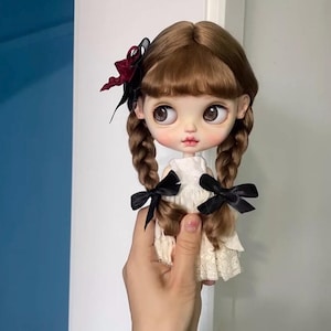 Puede incluir: Una muñeca con el pelo castaño que lleva un vestido blanco y lazos negros en el pelo. La muñeca tiene grandes ojos azules y una sonrisa pintada.