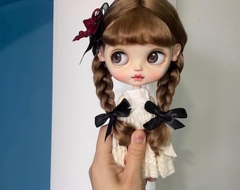 May include: A brown haired doll with a white dress and black bows in her hair. The doll has large blue eyes and a painted smile.