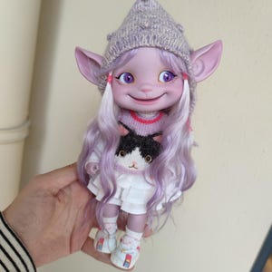 Puede incluir: Una muñeca morada con el pelo largo y rizado morado, que lleva un vestido blanco, un suéter rosa y morado con un diseño de gato blanco y negro, y un gorro de punto morado. La muñeca tiene ojos grandes y morados y una amplia sonrisa.