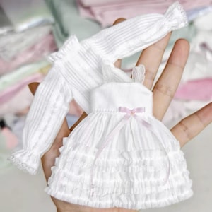 Op de afbeelding: Een witte poppenjurk met lange mouwen, een rok met ruches en een klein lichtroze strikje in de taille. De jurk heeft een geribbelde textuur op het lijfje en de mouwen, met een delicaat kanten randje bij de manchetten en de halslijn.