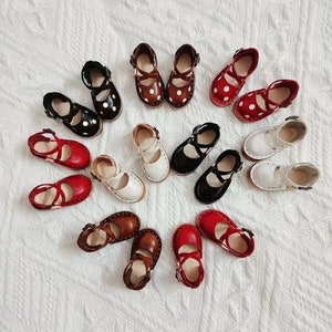 Può includere: Una collezione di 15 paia di scarpe in miniatura di vari colori, tra cui rosso, bianco, nero e marrone. Le scarpe hanno un design semplice con un cinturino sopra e una chiusura a fibbia. Alcune delle scarpe hanno pois bianchi.