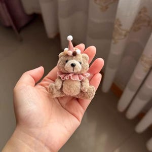 May include: A small, fuzzy, light brown teddy bear figurine wearing a dusty pink party hat topped with a white pom-pom and a matching dusty pink ruffled collar. The bear has dark eyes, a dark nose, and a soft, textured plush appearance.