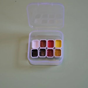 Op de afbeelding: Een transparante plastic doos met negen aquarelverfpannetjes. De pannetjes zijn gerangschikt in drie rijen van drie. De kleuren zijn wit, bruin, zwart, rood, oranje, geel en een lichtere bruintint.