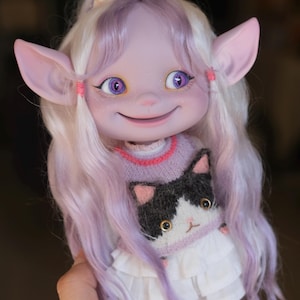 May include: A purple and white doll with large ears and long, wavy hair. The doll is wearing a pink and white sweater with a black and white cat design and a white skirt with ruffles.