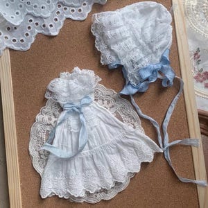 Peut inclure: Un ensemble robe et bonnet en broderie anglaise blanche avec des rubans bleu clair, présenté sur un tableau en liège. La robe a un col et une jupe à volants, le bonnet est bordé de bleu. L'ensemble est encadré.