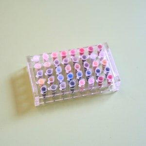May include: A clear plastic tray containing rows of small, colourful vials. The vials are filled with various shades of pink, red, blue, green, and brown.