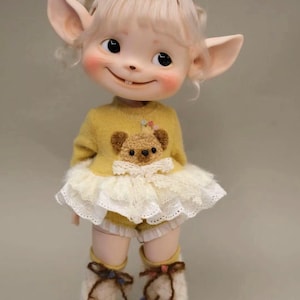 Puede incluir: Una pequeña muñeca con orejas grandes, cabello rubio y un suéter amarillo con un diseño de oso de peluche marrón. La muñeca lleva una falda de encaje blanco y polainas de lana blanca con lazos marrones.