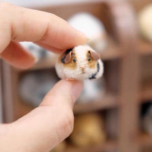 Pode incluir: Uma miniatura de porquinho-da-índia em feltro com pelagem branca, castanha e preta, pequenas orelhas escuras, nariz cor-de-rosa e olhos de contas pretas. A pequena escultura de animal com textura macia é segurada entre dois dedos.