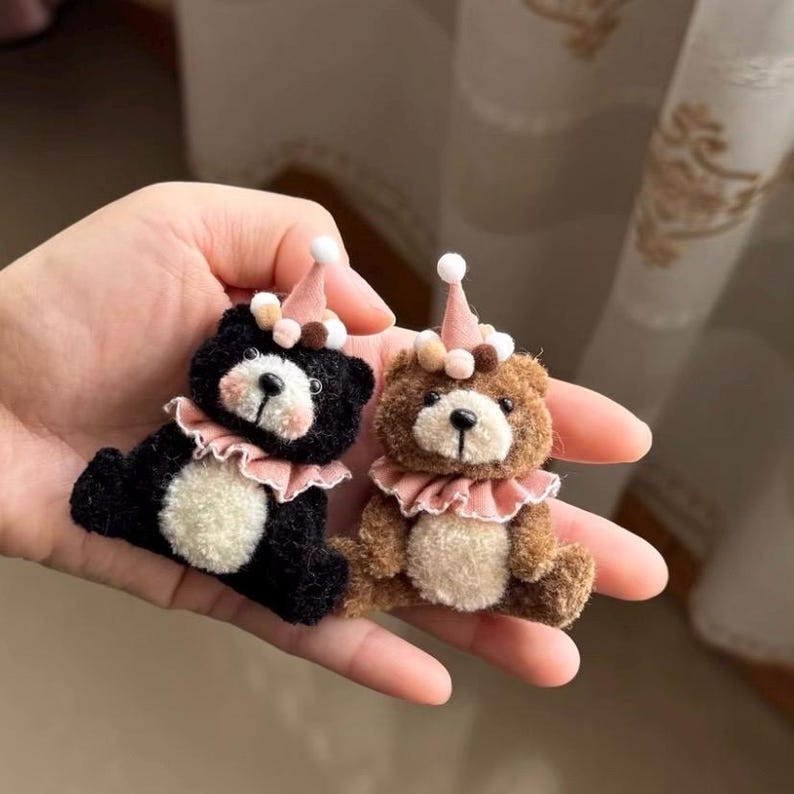 May include: Two small, fuzzy bear figurines, one black and one brown, each featuring a cream-coloured belly, a pink ruffled collar, and a matching pink party hat topped with a white pom-pom. The bears have dark eyes and stitched noses.