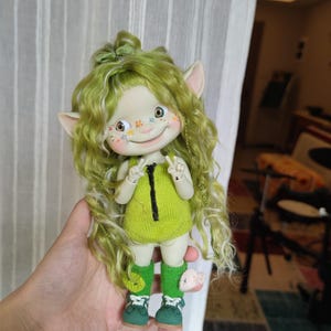 Puede incluir: Una muñeca verde y blanca con el pelo rizado, que lleva un mono de punto verde, calcetines verdes y zapatos verdes. La muñeca tiene orejas grandes y una cara pintada con flores.