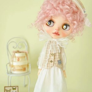 K&ouml;nnte beinhalten: Eine Puppe mit pinkfarbenen Locken, die ein wei&szlig;es Kleid, eine braune und wei&szlig;e karierte Weste und einen wei&szlig;en Hut mit blauem Besatz tr&auml;gt. Die Puppe schaut nach oben und hat blaue Augen.