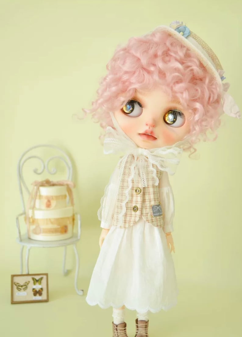 K&ouml;nnte beinhalten: Eine Puppe mit pinkfarbenen Locken, die ein wei&szlig;es Kleid, eine braune und wei&szlig;e karierte Weste und einen wei&szlig;en Hut mit blauem Besatz tr&auml;gt. Die Puppe schaut nach oben und hat blaue Augen.