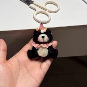 May include: A small, fuzzy black bear figurine with a cream-coloured belly and muzzle. The bear wears a pink party hat topped with a white pom-pom and a pink ruffled collar. The bear has small, round, multi-coloured pom-poms around the hat base.