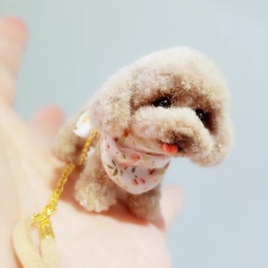 Puede incluir: Un pequeño juguete de perro, marrón y esponjoso, con un pañuelo floral rosa y blanco. El juguete lleva una cadena dorada.