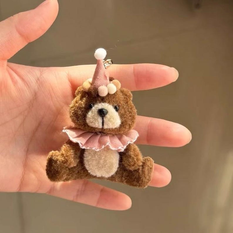 May include: A small brown fuzzy bear keyring featuring a light beige belly and muzzle. The bear wears a dusty pink party hat topped with a white pom-pom and a matching dusty pink ruffled collar around its neck, attached to a silver metal ring.