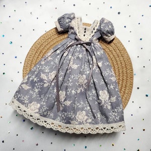 Pode incluir: Um vestido azul em miniatura com um padrão floral e acabamento em renda branca. O vestido apresenta mangas curtas bufantes, um decote com acabamento em renda e um laço na cintura. É exibido em um tapete circular tecido.