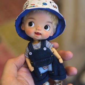 May include: A doll wearing a blue and white patterned bucket hat and blue dungarees. The doll has blonde hair, rosy cheeks, and is wearing blue and white shoes. The doll's dungarees have a small patch with a bear on it.