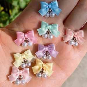 Pode incluir: Uma coleção de laços de cetim coloridos com detalhes de pérolas. Os laços, em tons pastel como azul, rosa, amarelo e lilás, têm duas pérolas penduradas presas com um acessório de metal prateado, criando um visual delicado.