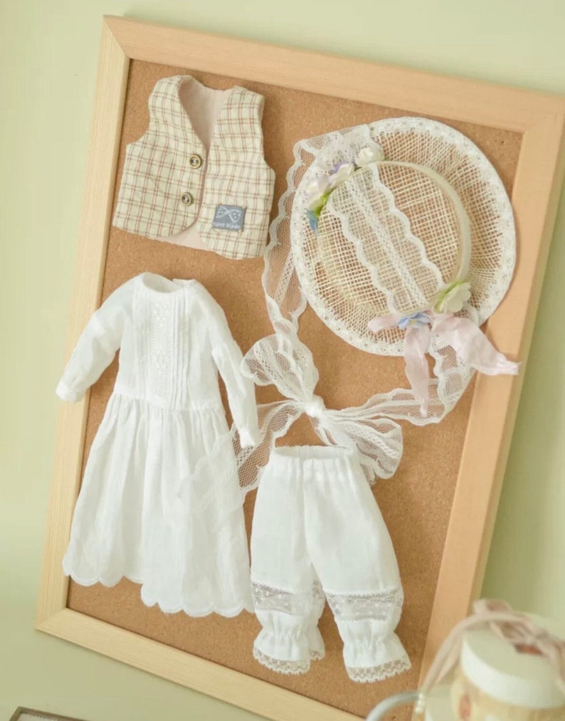 K&ouml;nnte beinhalten: Ein Set Puppenkleidung, das auf einer Korktafel ausgestellt ist. Das Set beinhaltet ein wei&szlig;es Kleid mit Spitzenbesatz, eine wei&szlig;e Hose mit Spitzenbesatz, eine karierte Weste und einen Strohhut mit Spitzen- und Blumenapplikationen.