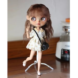Puede incluir: Una muñeca personalizada con cabello castaño y ojos grandes y expresivos. La muñeca viste un vestido con estampado floral con cuello Peter Pan, zapatos blancos y un bolso bandolera negro. Está posada sobre un soporte blanco.