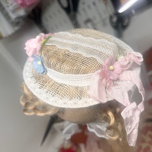 K&ouml;nnte beinhalten: Ein wei&szlig;er Spitzenhut mit rosa und blauen Stoffblumen. Der Hut hat ein rosa Band, das um ihn gebunden ist.