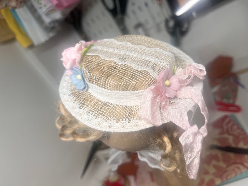 K&ouml;nnte beinhalten: Ein wei&szlig;er Spitzenhut mit rosa und blauen Stoffblumen. Der Hut hat ein rosa Band, das um ihn gebunden ist.
