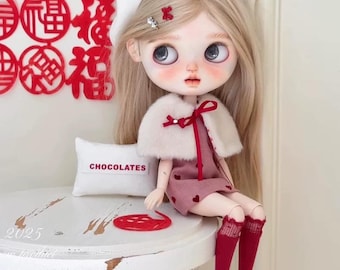 May include: A doll with long blonde hair, wearing a pink dress with heart patterns, a white beret, and red socks. The doll is seated on a white table with a small pillow that says "CHOCOLATES". Decorative red Chinese characters are on the wall.