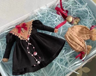 May include: A black dress with a lace collar and a red ribbon. The dress has a beaded necklace and a cross on the collar. The dress is in a box with white tissue paper.