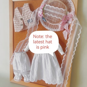 K&ouml;nnte beinhalten: Eine Korktafel mit einem wei&szlig;en Kleid, einer wei&szlig;en Hose mit Spitzenbesatz, einer karierten Weste und einem wei&szlig;en Spitzenhut mit rosa Blumen. Hinweis: Der neueste Hut ist rosa.