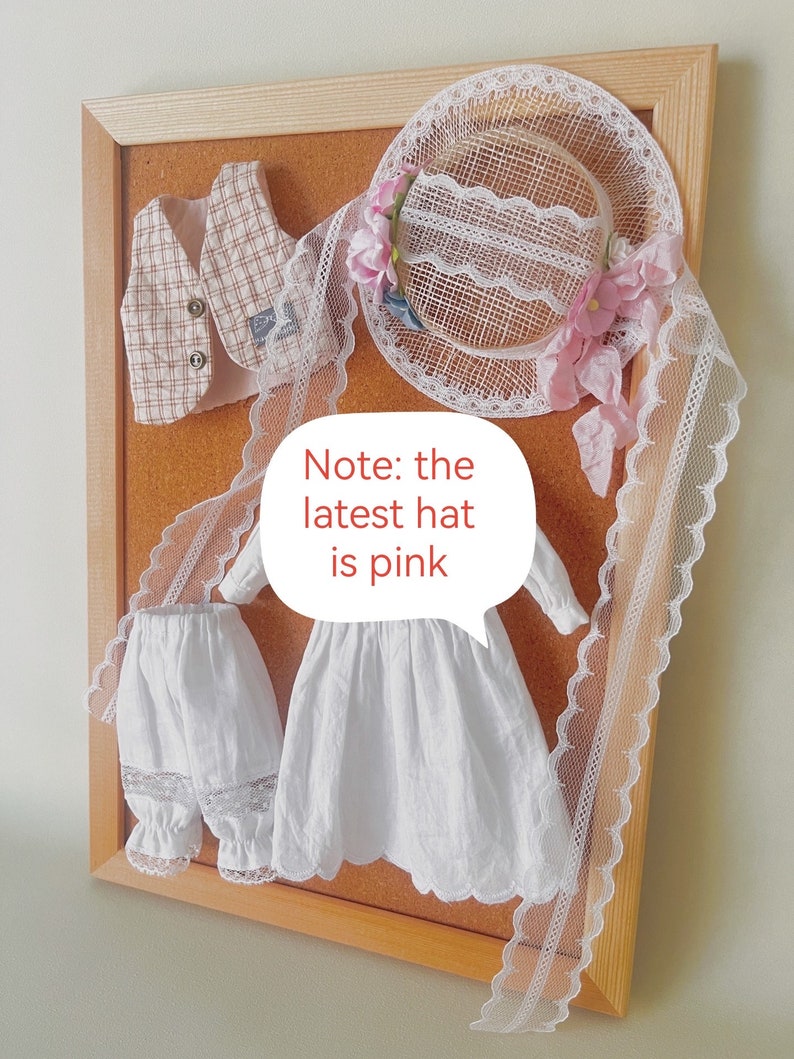 K&ouml;nnte beinhalten: Eine Korktafel mit einem wei&szlig;en Kleid, einer wei&szlig;en Hose mit Spitzenbesatz, einer karierten Weste und einem wei&szlig;en Spitzenhut mit rosa Blumen. Hinweis: Der neueste Hut ist rosa.