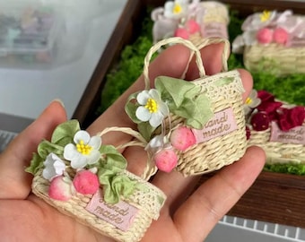 May include: Miniature woven baskets with floral and fruit embellishments. Each basket features a handle, lace trim, and a pink label that reads "hand made". The baskets are decorated with white flowers, green leaves, and pink fruit.