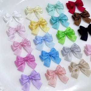 Può includere: Una collezione di piccoli fiocchi in tessuto in una varietà di colori tra cui bianco, giallo, azzurro, rosso, marrone, nero, verde, argento, rosa, viola e beige. Ogni fiocco presenta una superficie testurizzata ed è disposto a griglia.