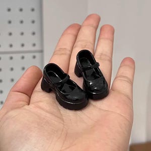 May include: A pair of miniature black Mary Jane shoes with a glossy finish. The shoes have a chunky heel and a strap across the top. These doll shoes are a classic style, perfect for a doll or collectible.