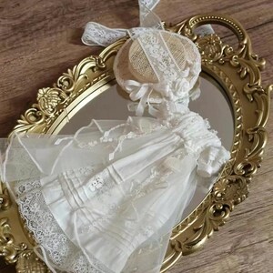 Peut inclure: Une robe de poupée blanche avec une bordure en dentelle et un bonnet assorti. La robe est présentée sur un miroir doré avec des détails ornés. Un ruban blanc est attaché au bonnet. La robe est en tissu délicat.