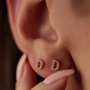 May include: Close-up of two small gold-coloured stud earrings, each shaped like the letter "D". The earrings are encrusted with small, clear gemstones. The earrings are in a person's ear.