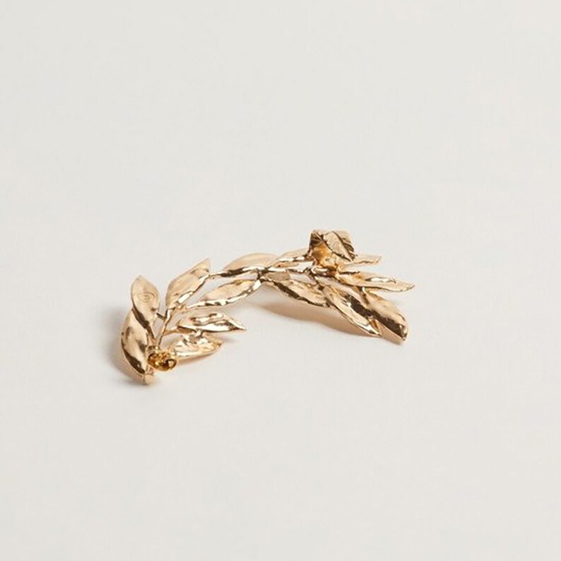 May include: A gold-colored leaf-shaped ear cuff. The ear cuff features a detailed design of leaves and stems, with a textured surface. The jewelry is set against a white background.