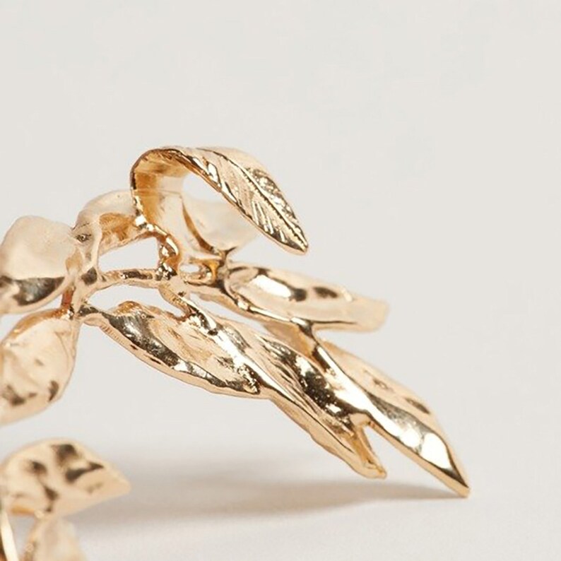 May include: Close-up of a gold-colored leaf-shaped jewelry piece. The leaves have detailed veining and a textured surface, with a bright, reflective finish. The design suggests a nature-inspired theme, possibly a brooch or earring.