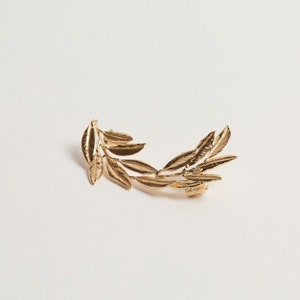 May include: Gold-colored ear cuff featuring a laurel leaf design. The ear cuff is made of metal and has a detailed, realistic leaf pattern. The jewelry piece is designed to be worn on the ear and adds a touch of elegance.