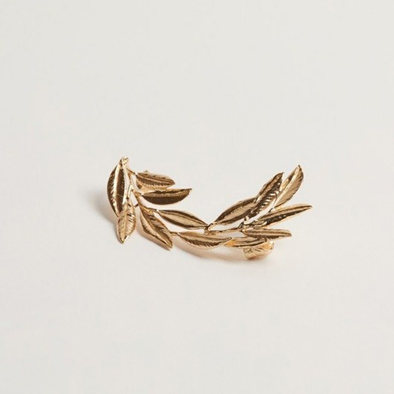 May include: Gold-colored ear cuff featuring a laurel leaf design. The ear cuff is made of metal and has a detailed, realistic leaf pattern. The jewelry piece is designed to be worn on the ear and adds a touch of elegance.