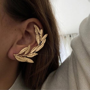 May include: Gold-colored ear cuff featuring a leaf design. The ear cuff wraps around the ear, with multiple leaves extending along the ear's curve. The jewelry is made of a shiny, metallic material.