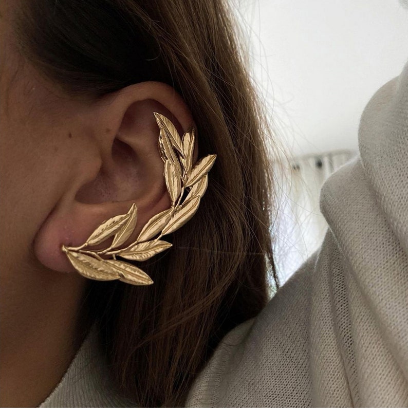 May include: Gold-colored ear cuff featuring a leaf design. The ear cuff wraps around the ear, with multiple leaves extending along the ear's curve. The jewelry is made of a shiny, metallic material.