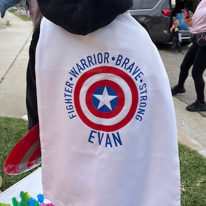 May include: White superhero cape with a red, white, and blue Captain America shield design. The cape has the words "Fighter, Warrior, Brave, Strong" around the shield and the name "Evan" below the shield.
