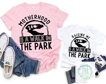 May include: Pink and white t-shirts with a black dinosaur skull graphic and the text "Motherhood is a walk in the park" and "Raising me is a walk in the park".