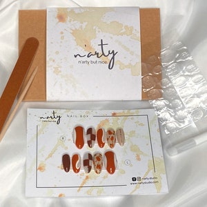 Op de afbeelding: Een set met opdrukbare nagels met een abstract ontwerp in bruin, wit en oranje. De set wordt geleverd in een doos met een nagelvijl, lijm en plakstrips. De doos heeft de tekst "n'arty n'arty but nice." erop.