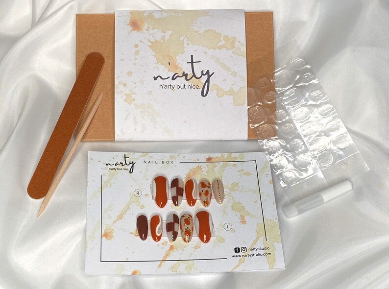 Op de afbeelding: Een set met opdrukbare nagels met een abstract ontwerp in bruin, wit en oranje. De set wordt geleverd in een doos met een nagelvijl, lijm en plakstrips. De doos heeft de tekst "n'arty n'arty but nice." erop.