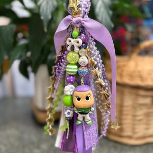 May include: A decorative keychain featuring a Buzz Lightyear figurine. The keychain is adorned with purple ribbons, beads, and gold star accents. A green loop and black clasp are at the top.